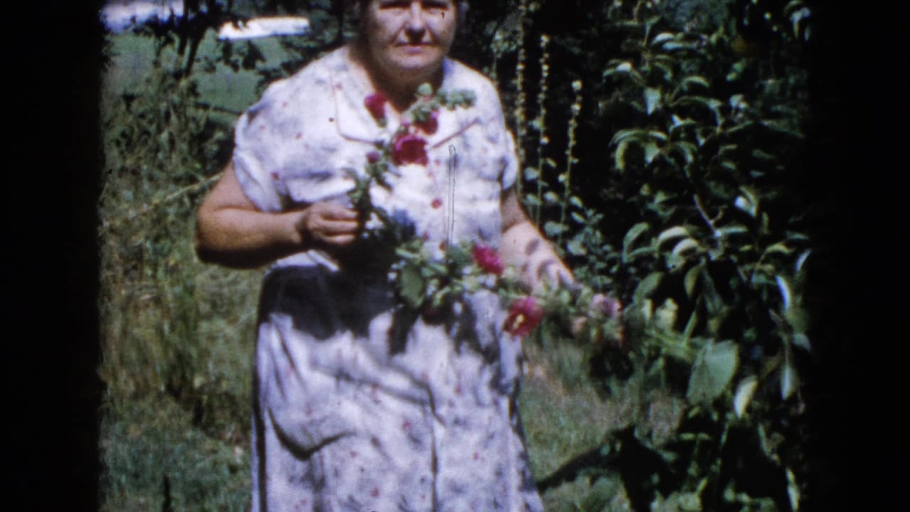 Woman faces the camera while holding several red flowers in a garden.