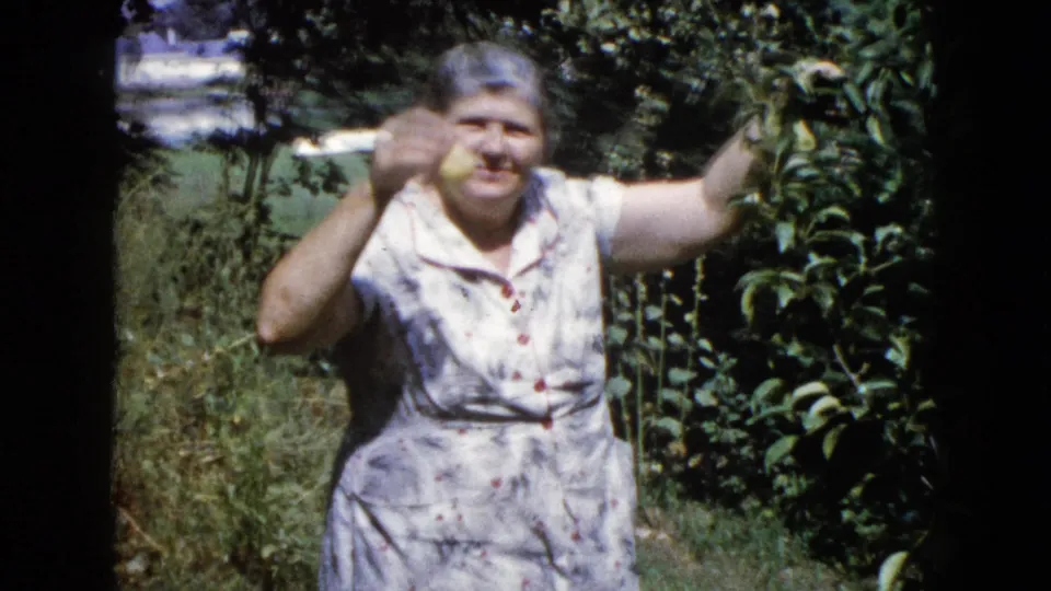 In 1960 Wauconda, Illinois home-movie footage, an older woman smiles while holding fruit near a tree in a sunlit garden.