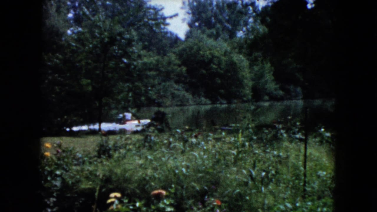 1960 Wauconda home-movie frame showing dense garden growth, trees, calm water, and a small boat near the opposite bank.
