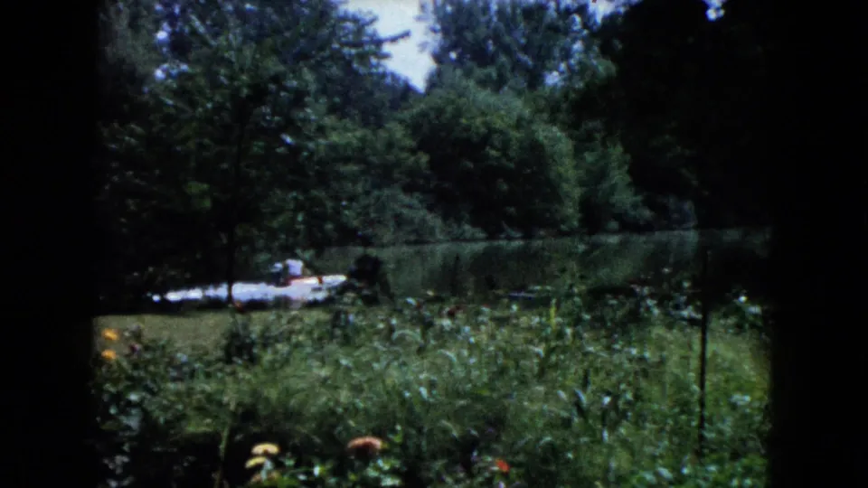 1960 Wauconda home-movie frame showing dense garden growth, trees, calm water, and a small boat near the opposite bank.
