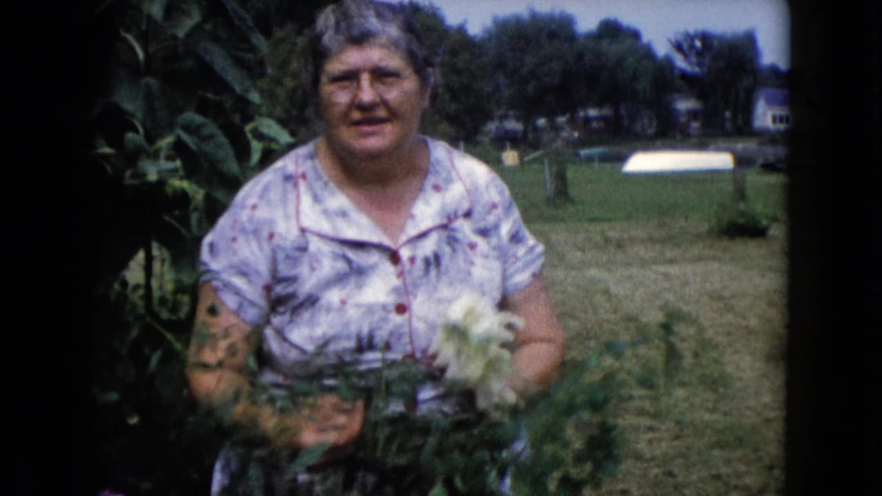 Older woman smiles toward the camera while holding white flowers near garden plants.
