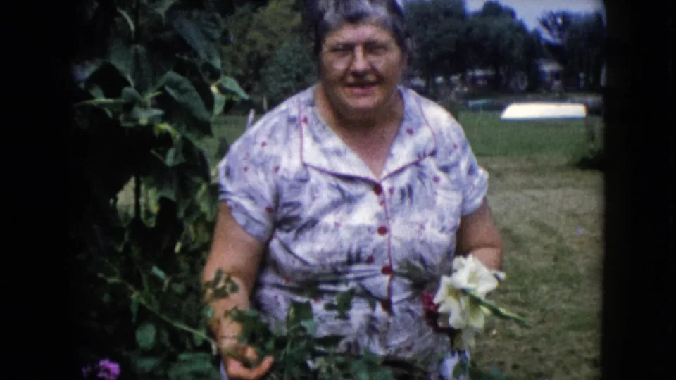 The woman in a floral dress holds white flowers and cut greenery near the garden in Wauconda, Illinois home-movie footage.