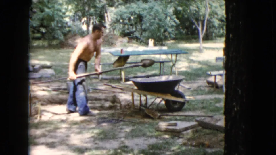 A shirtless young man in jeans pushes a wheelbarrow across a Wauconda backyard with trees and a picnic table visible behind him.