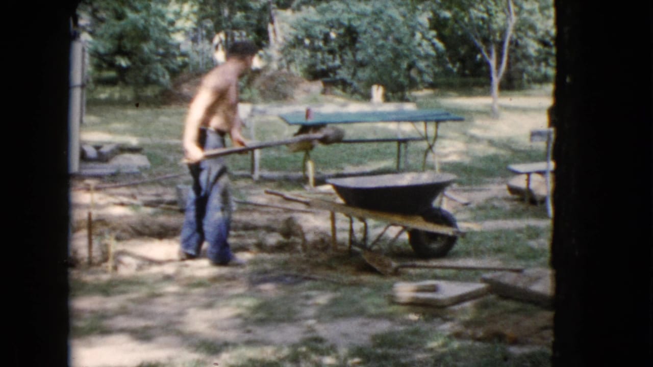 Shirtless man shoveling near a wheelbarrow and green picnic table in a shaded yard.
