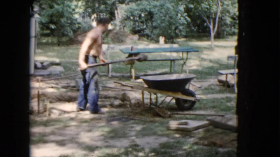 In Wauconda, Illinois, a shirtless man in jeans works with a shovel beside a wheelbarrow and green picnic table in a tree-lined yard.