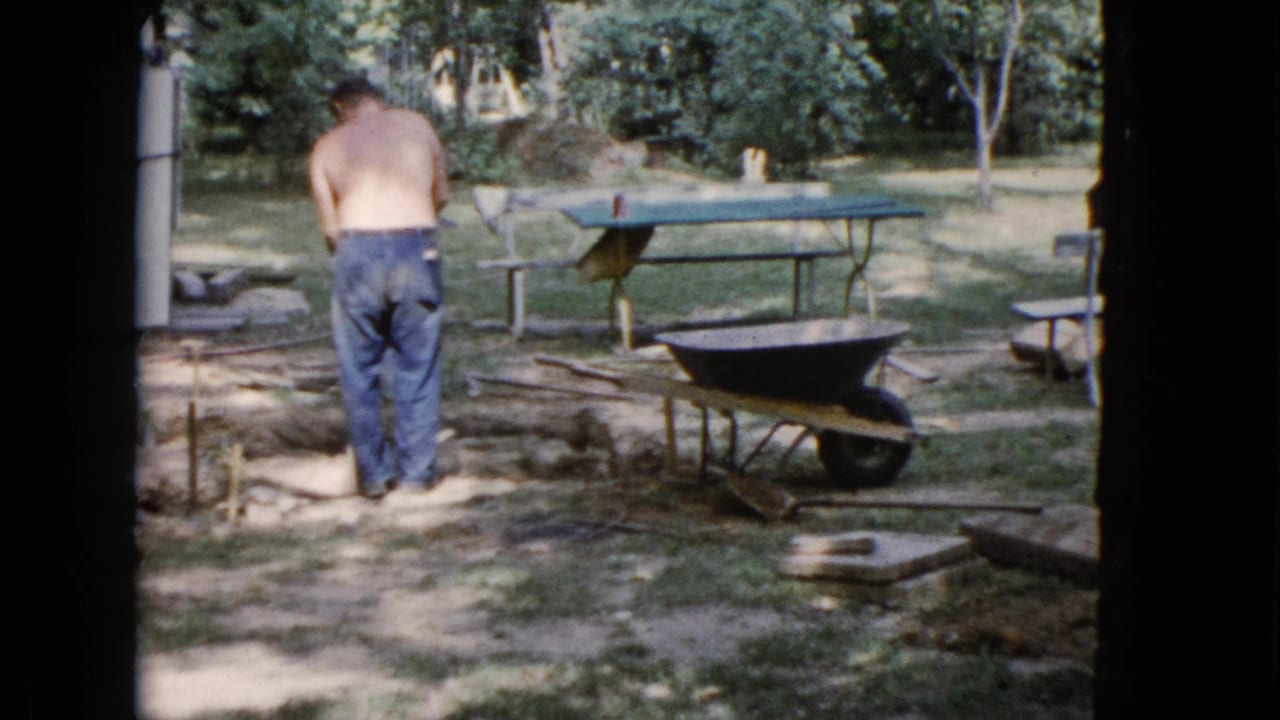 The man is filmed from behind while shoveling in the yard, with a wheelbarrow, picnic table, trees, and loose materials nearby.