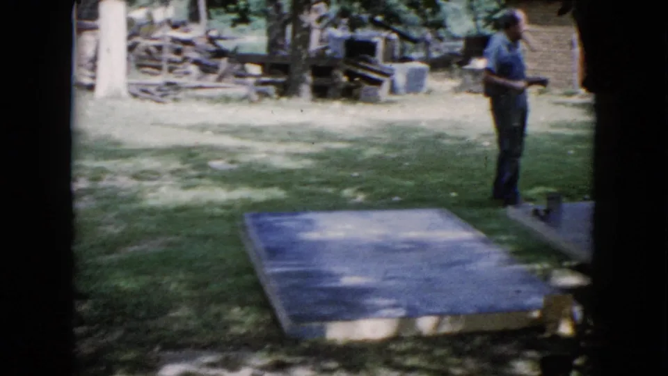 A man stands in the shaded yard near a flat platform or boards, with fencing, trees, and scattered outdoor materials visible behind him.