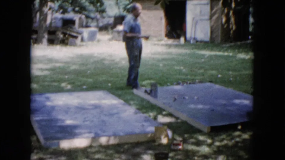 The man remains near the slabs in the 1960 Wauconda backyard, with trees and a brick outbuilding behind him.
