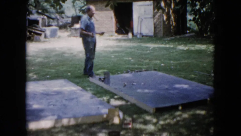 A man in a blue shirt looks down toward the slab area while holding a measuring tool in the Illinois backyard scene.