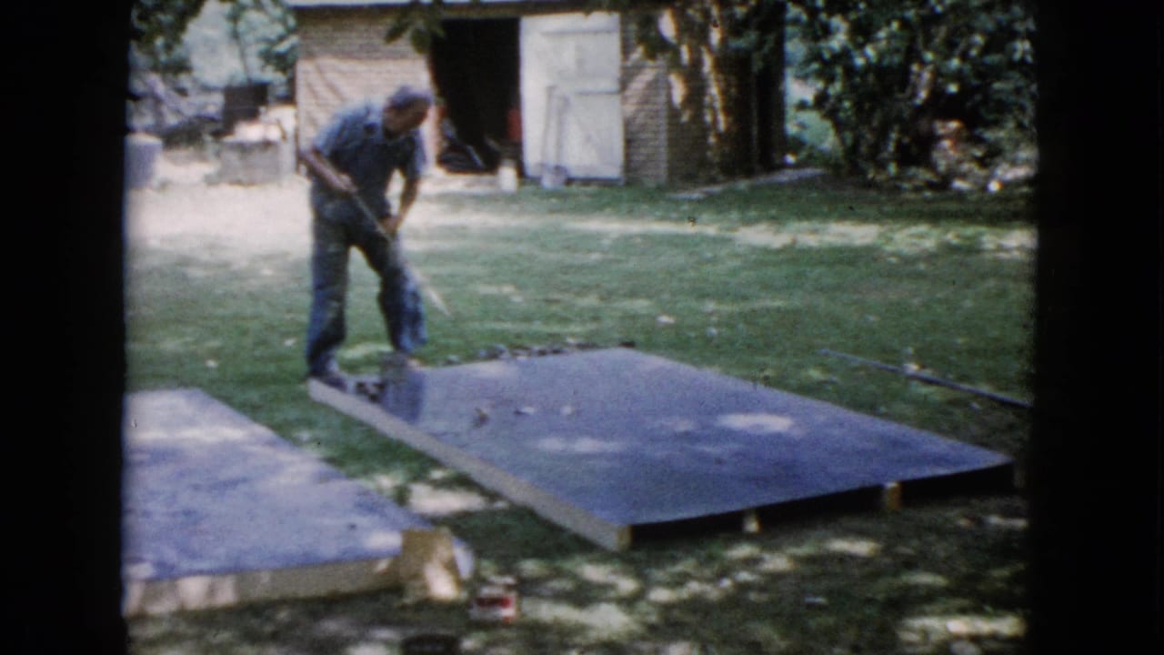 Man in a blue shirt works over a large slab on grass near a shed.
