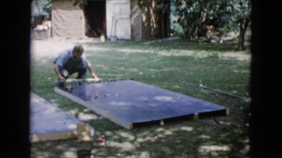 A man kneels beside a large slab in a Wauconda backyard, with a shed, trees, and lawn visible in the 1960 8mm frame.