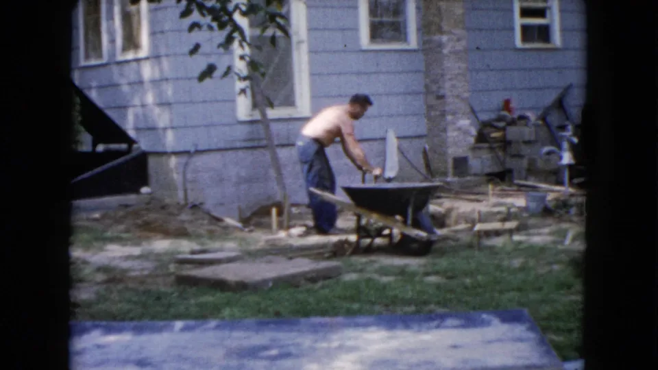 A shirtless man shovels near a wheelbarrow beside a light blue house in 1960 Wauconda backyard home-movie footage.