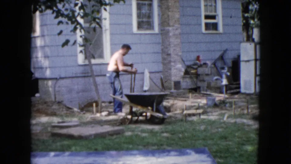 The 1960 Wauconda frame shows a shirtless man standing at a wheelbarrow near a shovel, house wall, grass, and scattered materials.