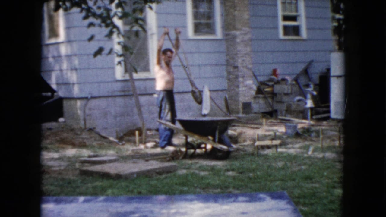 Shirtless man holds a shovel beside a wheelbarrow in a backyard near a blue house.