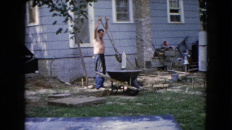 In Wauconda, Illinois in 1960, a shirtless man stands with a shovel near a wheelbarrow outside a light blue house.
