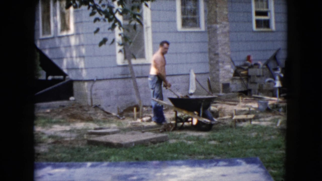 Man walks or turns near a wheelbarrow and shovel in a residential yard.