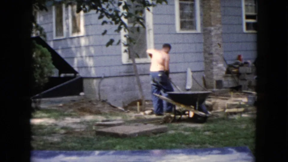 The man faces away from the camera beside the wheelbarrow, framed by a tree trunk, house siding, and scattered yard materials.