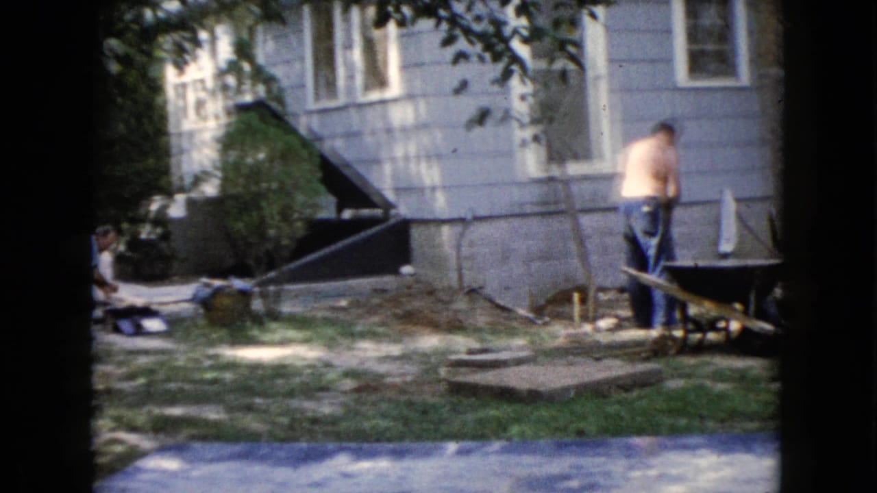 1960 Wauconda home-movie frame showing two men doing yard work beside a light-sided house, with a wheelbarrow, shovel, grass, and trees visible.