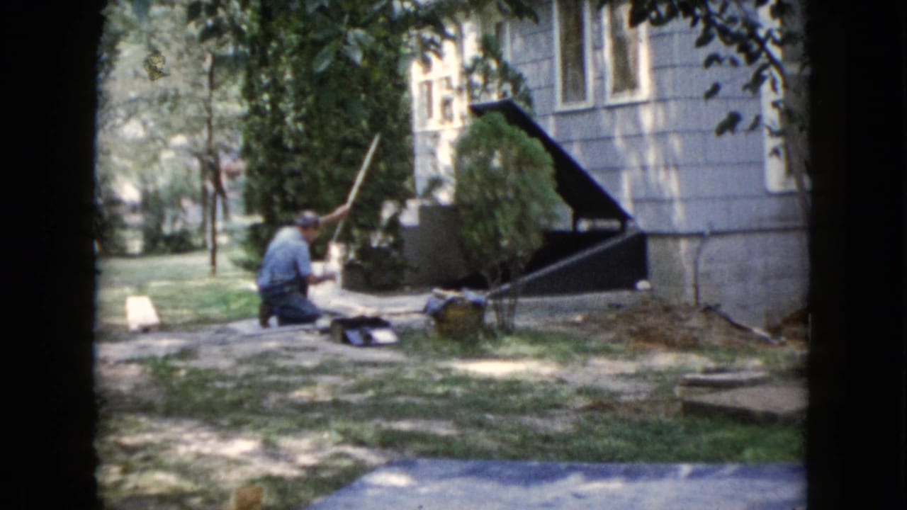 1960 Wauconda yard scene with a man kneeling near the ground beside a house exterior, shaded lawn, shrubs, and a walkway.