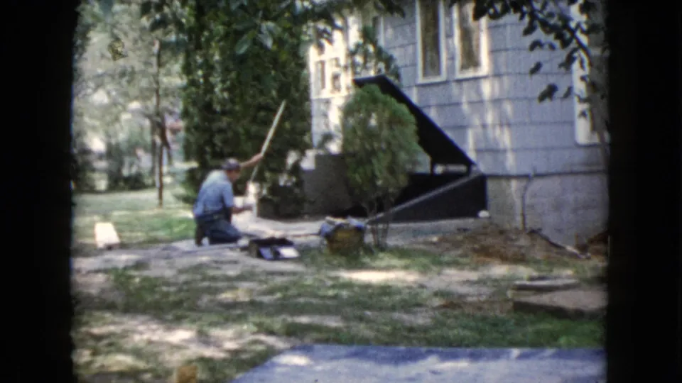 1960 Wauconda yard scene with a man kneeling near the ground beside a house exterior, shaded lawn, shrubs, and a walkway.