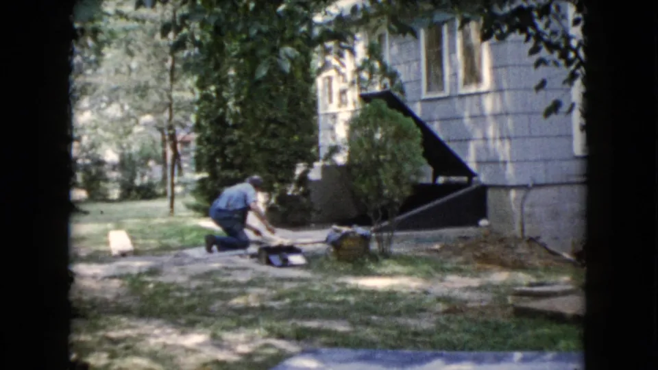 1960 Wauconda home-movie frame of a kneeling man doing ground-level yard work beside a light-sided residential house and lawn.