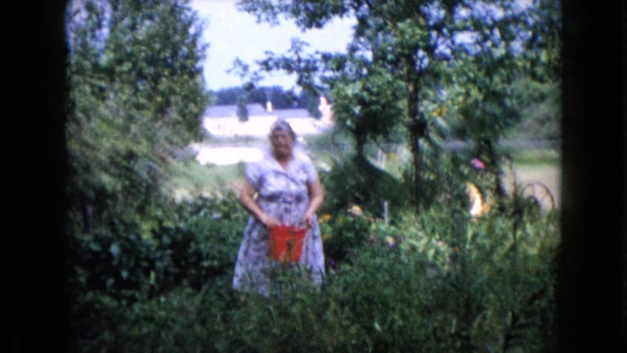 Woman in a patterned dress stands in a green garden holding a red bucket.