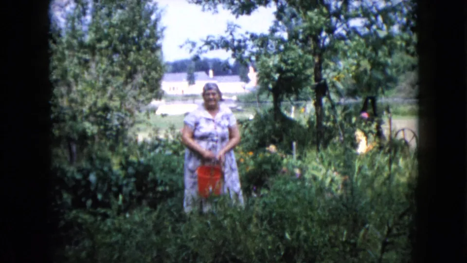 A woman in a patterned dress faces the camera in a Wauconda garden, with a red bucket and distant house visible.