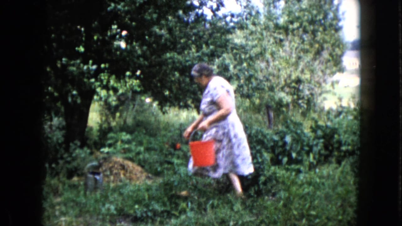 Woman carries a red bucket beside tall garden plants and trees.