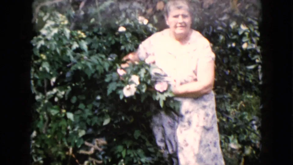 Older woman in a pale loose gown stands in a leafy Wauconda garden, holding light-colored flowers on 8mm home-movie film from 1960.