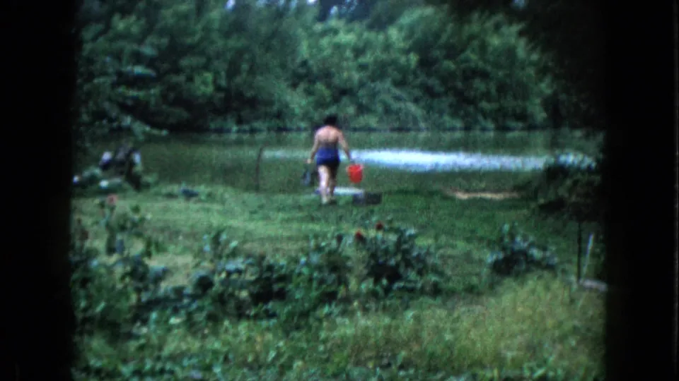 1960 Wauconda yard frame showing a younger woman in a blue bathing suit walking toward a pond with a red bucket.