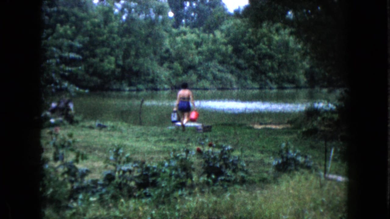 In Wauconda, Illinois, a woman in a swimsuit crosses a grassy yard toward a pond with a red bucket and dark container.