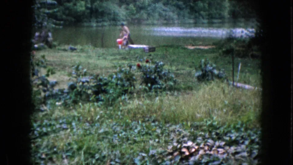 In the 1960 Wauconda backyard pond scene, the woman stands near the red bucket with water and trees behind her.