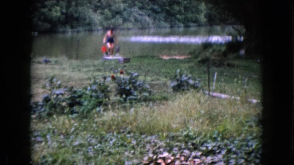 The woman stands near the Wauconda pond edge with a red bucket and low container, surrounded by grass, water, and trees.