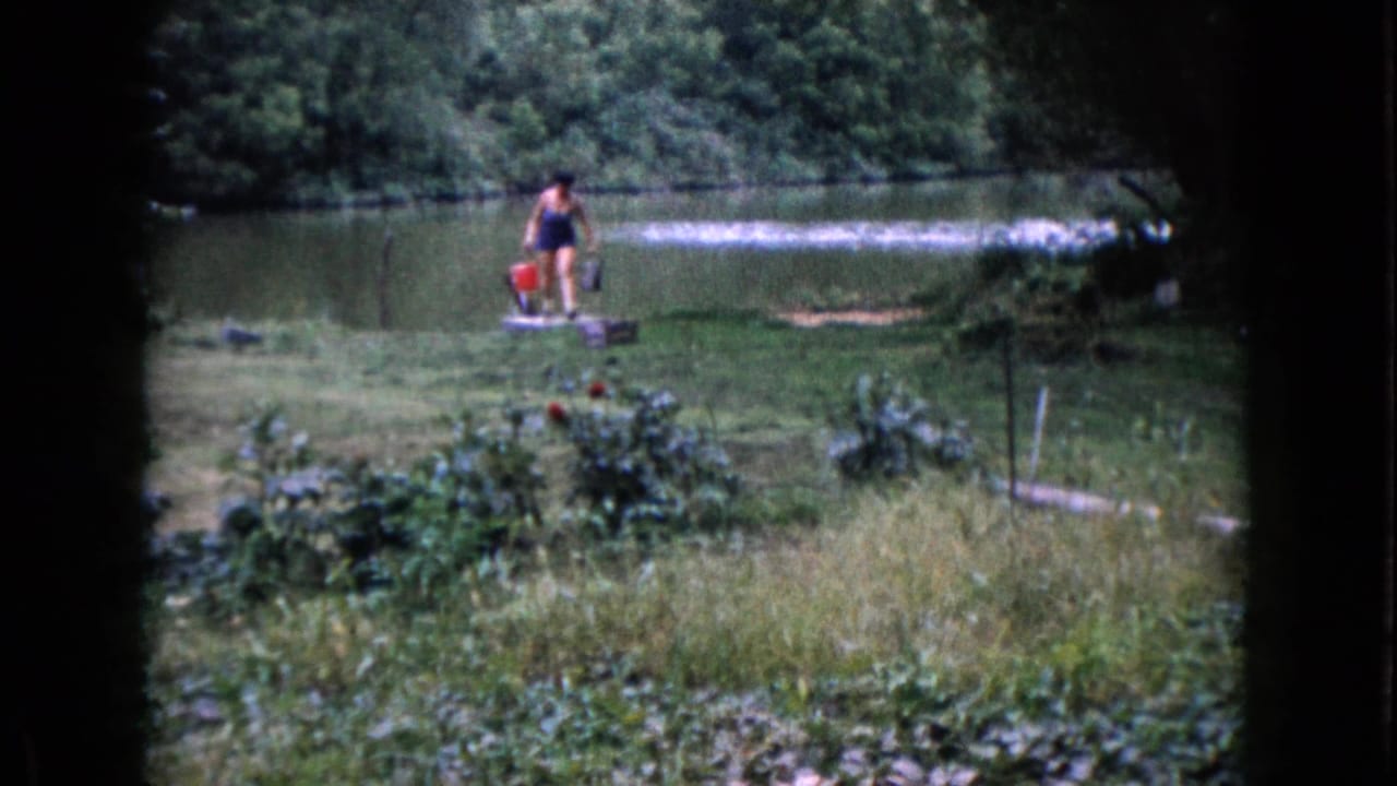 In 1960 Wauconda, Illinois footage, a woman in a dark swimsuit carries buckets across a grassy lakeshore.