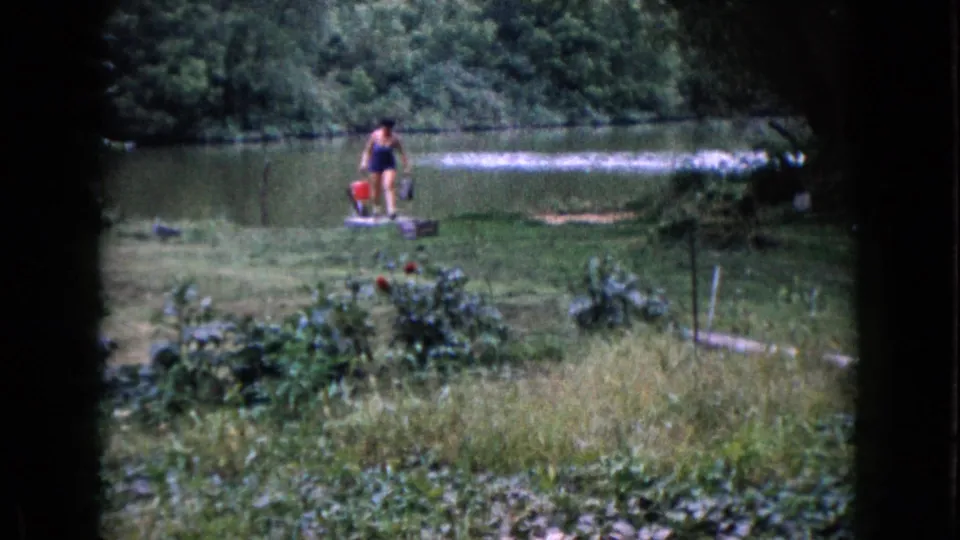 In 1960 Wauconda, Illinois footage, a woman in a dark swimsuit carries buckets across a grassy lakeshore.
