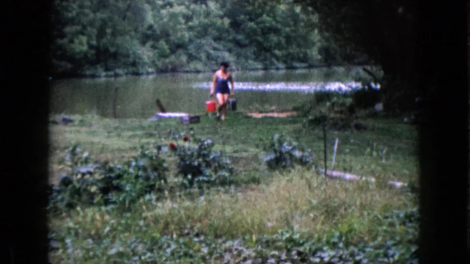 A 1960 home-movie frame shows the woman crossing the grassy shore with buckets, backed by lake water and trees.
