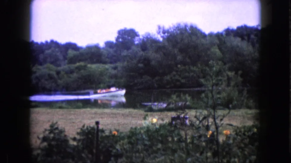 Small boat carrying people, some in bright life jackets, passes along a wooded lake in Wauconda, Illinois in 1960 footage.