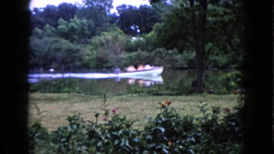 A fixed 8mm view shows the white boat near the wooded bank in Wauconda, Illinois, with foreground garden plants and grass.