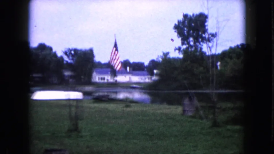 A Wauconda lakeside view shows an American flag near the water, with a distant house, trees, and small boats.