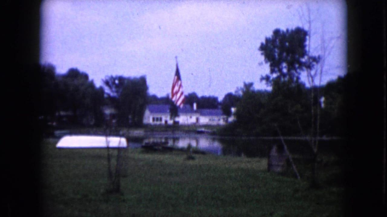 1960 Wauconda, Illinois 8mm frame showing an American flag by a lawn, white house, pond, trees, and a small white boat.