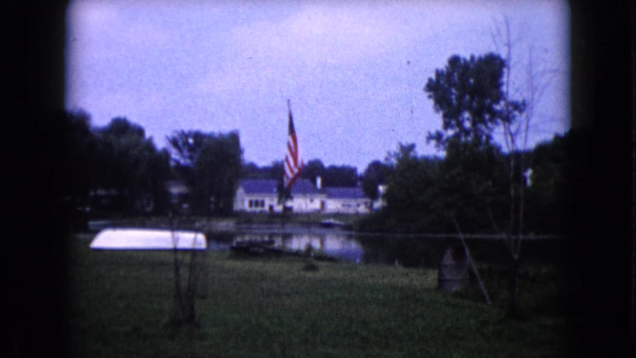 American flag beside water with a white boat on grass and houses behind trees.