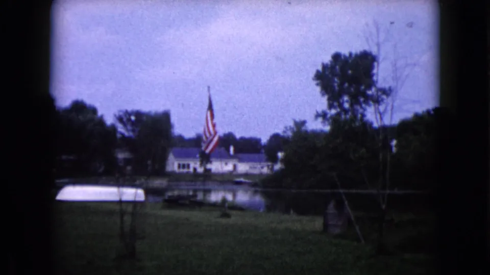 1960 Wauconda, Illinois 8mm view of an American flag near a pond-side yard, with a white boat, house, and trees visible.