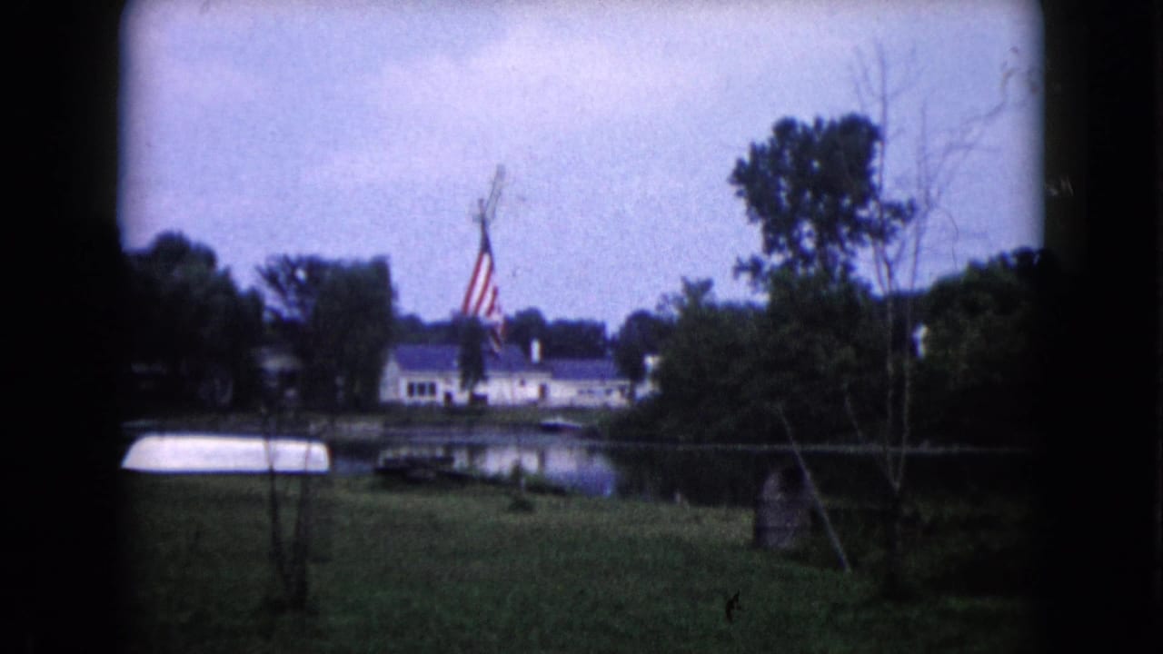 1960 Wauconda, Illinois home-movie frame showing a lakeside yard, American flag, dock, trees, and a white house across the water.