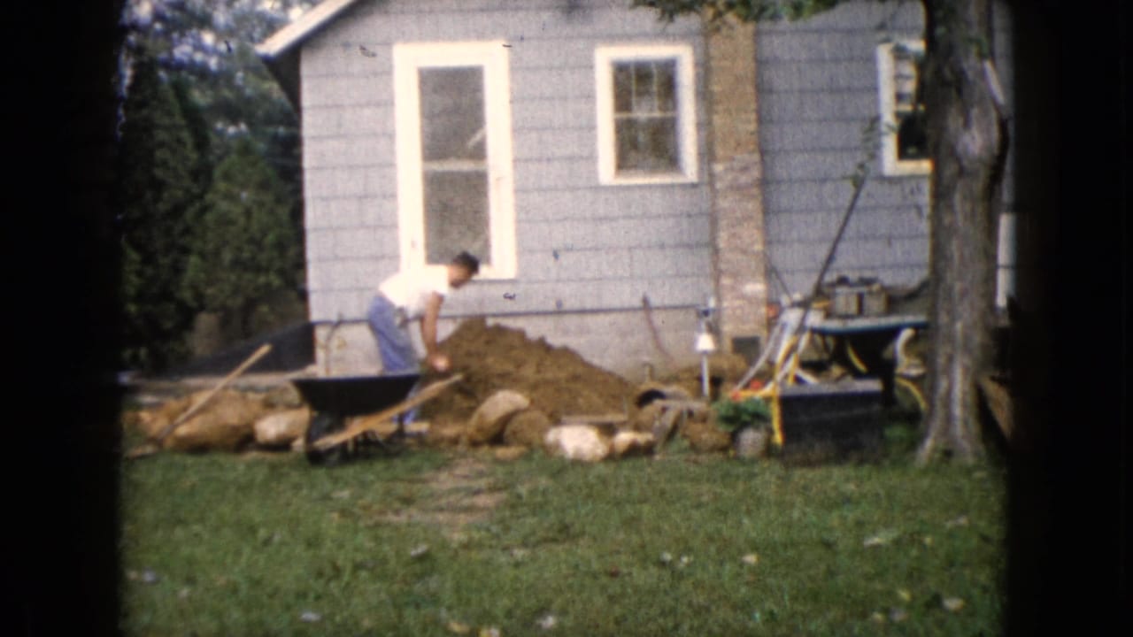 Man with a shovel working beside a wheelbarrow near a gray-sided house.