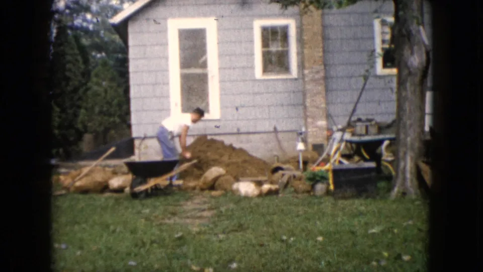 1960 Wauconda, Illinois home-movie frame of a man shoveling near a wheelbarrow, dirt pile, rocks, and gray-sided house.