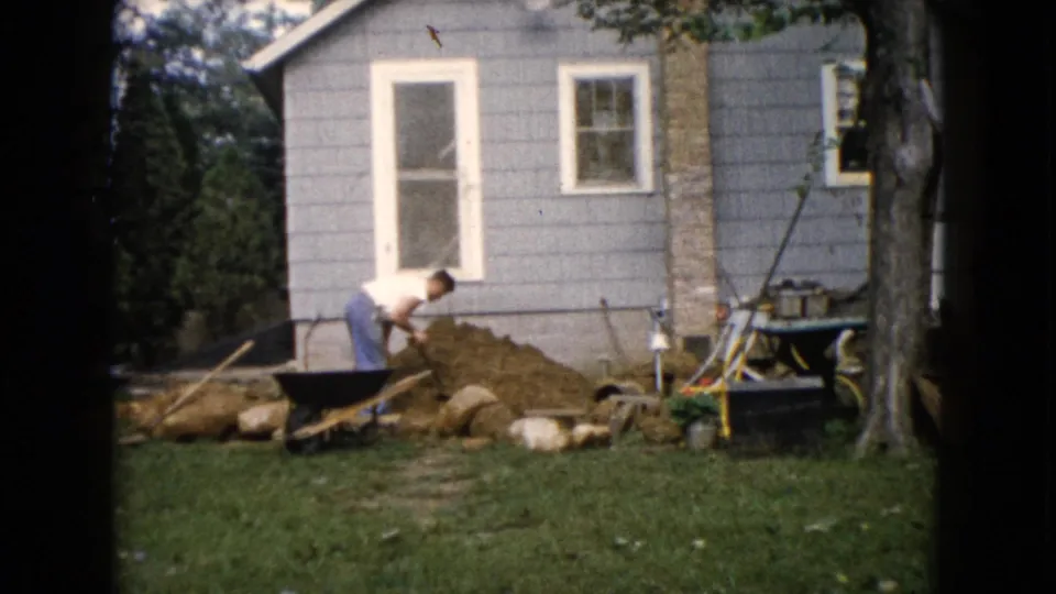 1960 Wauconda, Illinois home-movie frame showing a man bending to shovel soil beside rocks, a wheelbarrow, and a residential exterior.