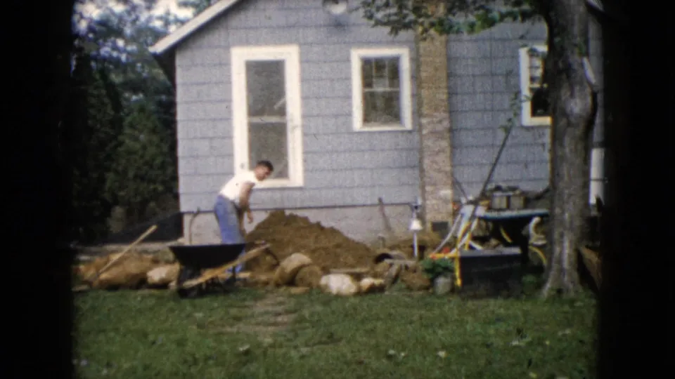 1960 Wauconda, Illinois home-movie frame of a man standing near yard work tools, a dirt pile, rocks, and a gray-sided house.