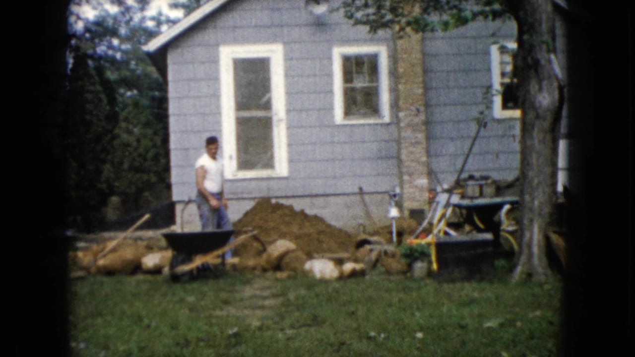 Man standing near a wheelbarrow and dirt pile beside a gray house.