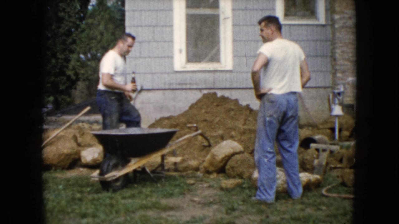 Two adult men in white shirts and jeans stand by a wheelbarrow and dirt pile outside a gray house in Wauconda, Illinois, 1960.