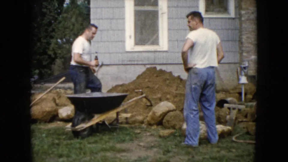 Two adult men in white shirts and jeans stand by a wheelbarrow and dirt pile outside a gray house in Wauconda, Illinois, 1960.